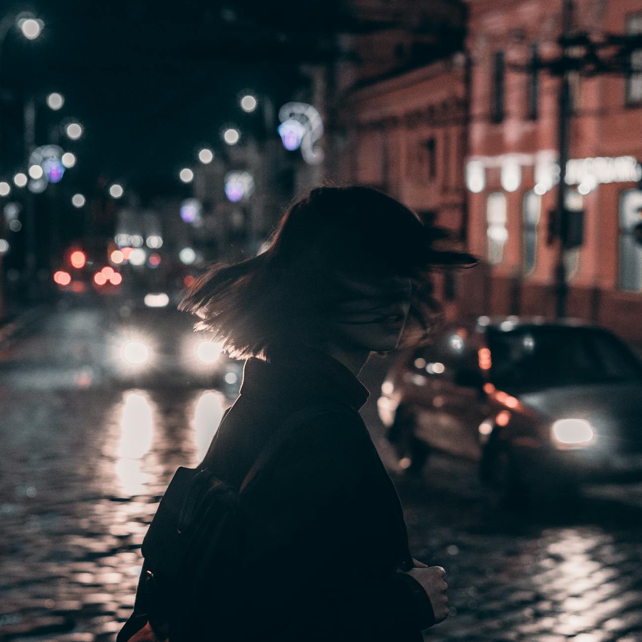 woman in black jacket carrying backpack with tousled hair walking on the street at night